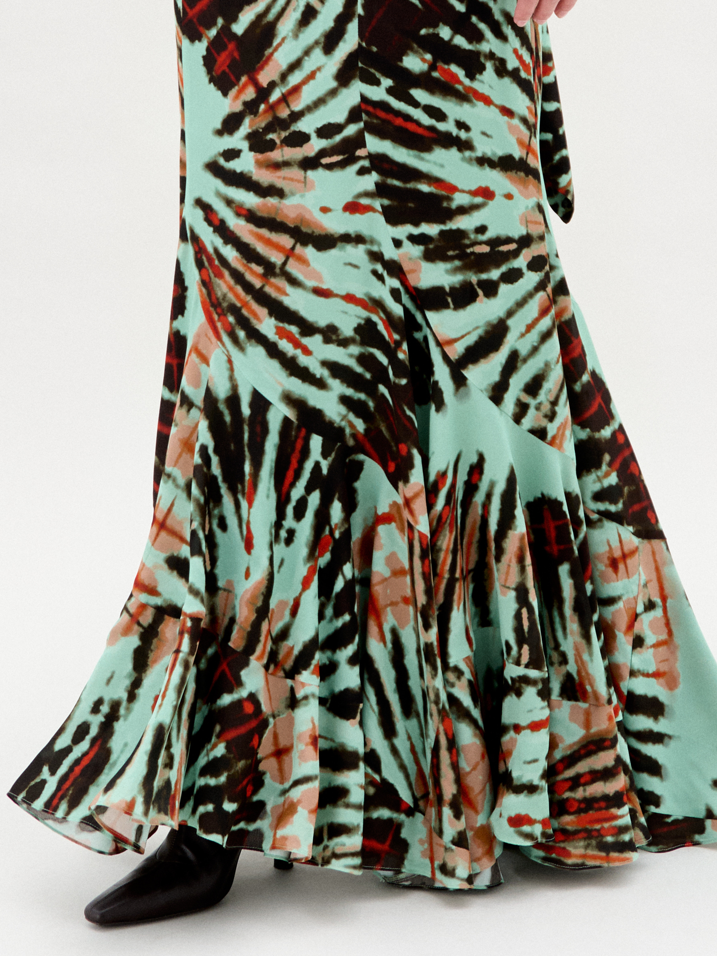 Against a plain white background, a person wears the Florina Skirt, crafted from luxurious Italian fabric. This long skirt showcases a vibrant tie-dye pattern with green, orange, and black hues cascading elegantly down its maxi length.