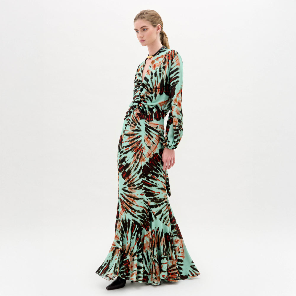 A woman poses against a white background in the SVT DESIGNS Florina Skirt, featuring a green, brown, and black abstract pattern with a mermaid silhouette and flared hem. She pairs it with a long-sleeve top, v-neckline, and black shoes.