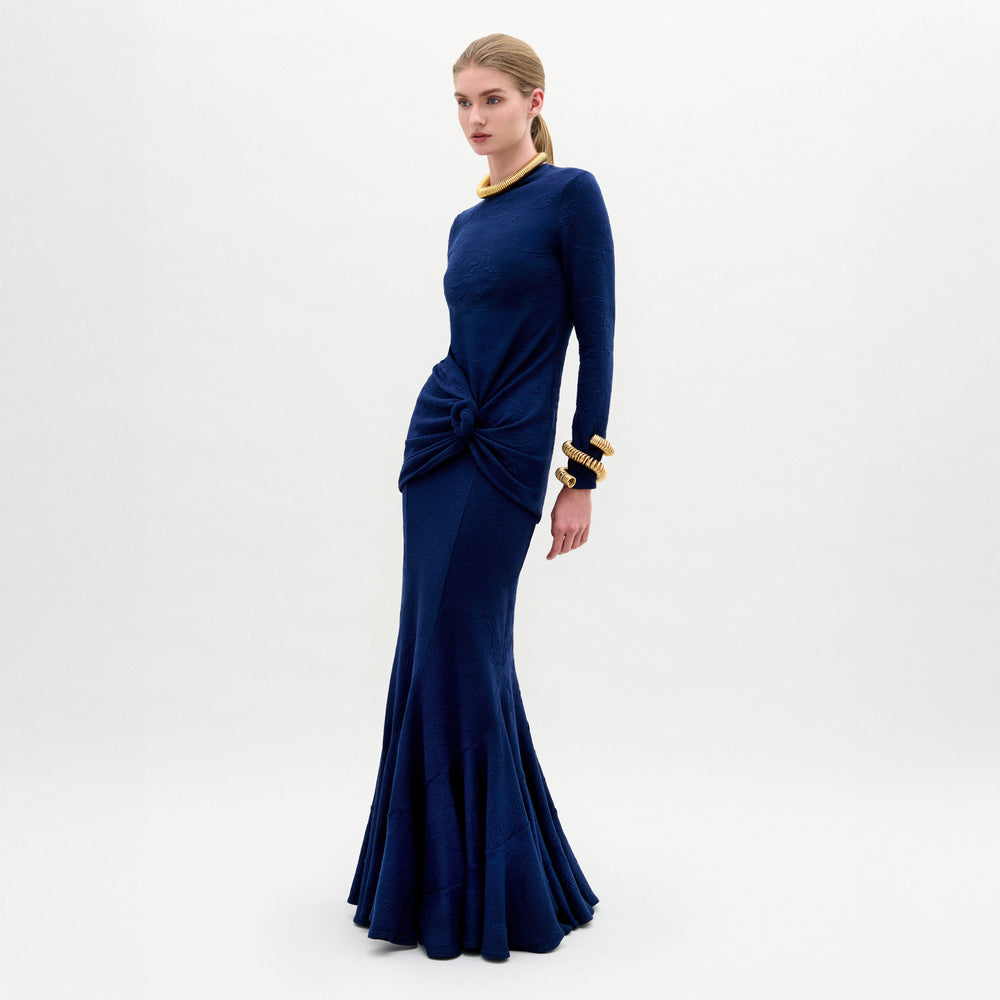 A woman stands against a white background wearing the SVT DESIGNS Florina Skirt, a long, dark blue fitted skirt in luxurious Italian fabric with a knot detail at the hips. She accessorizes with a gold necklace and bangles.