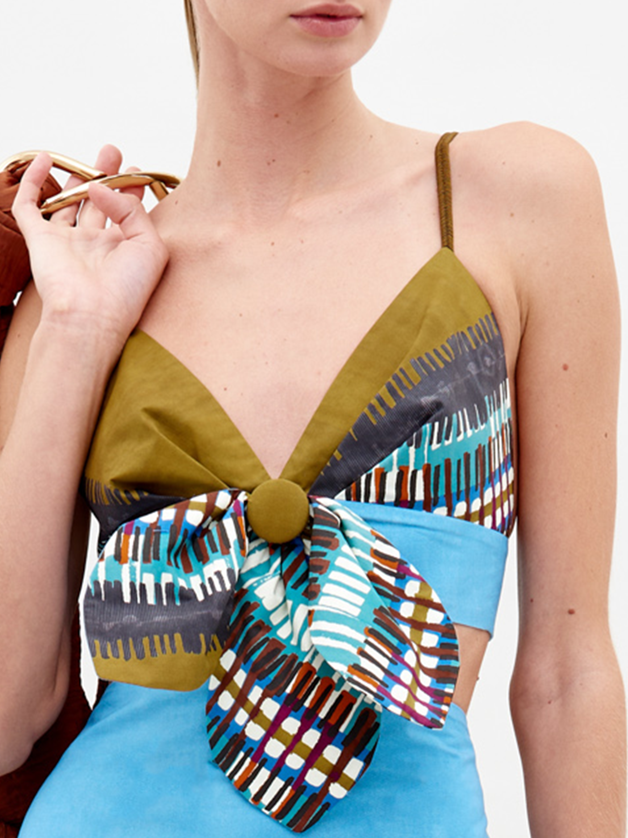 A person wearing the Flor Top by SVT DESIGNS—a sleeveless piece with olive green straps, a deep V neckline, a large front bow detail, and blue, brown, and white Italian-inspired geometric patterns.