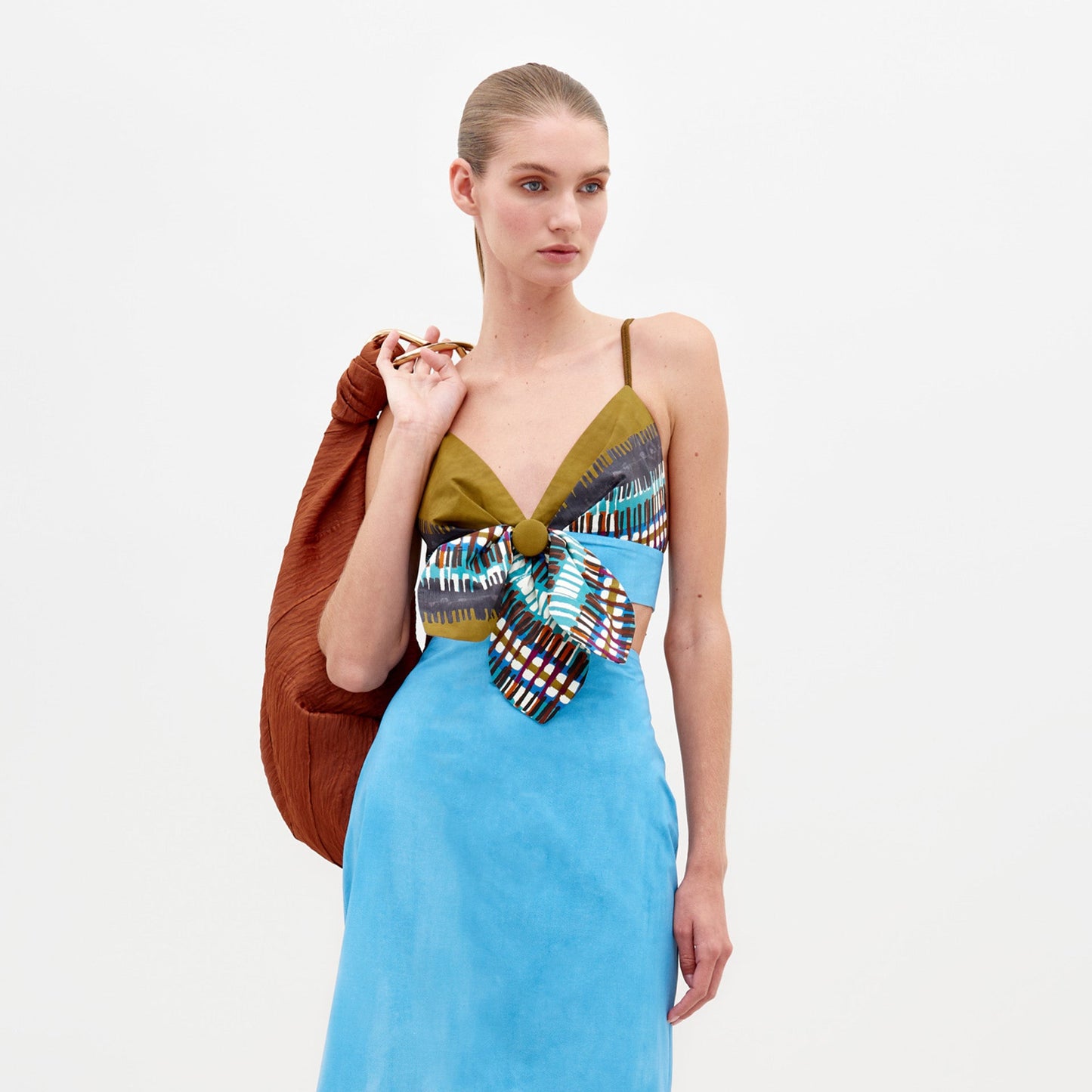 A woman stands against a plain white background wearing the Atira Skirt by SVT DESIGNS, a light blue Italian cotton piece with thin straps, a large multicolored bow at the front, and a brown textured accessory over one shoulder.