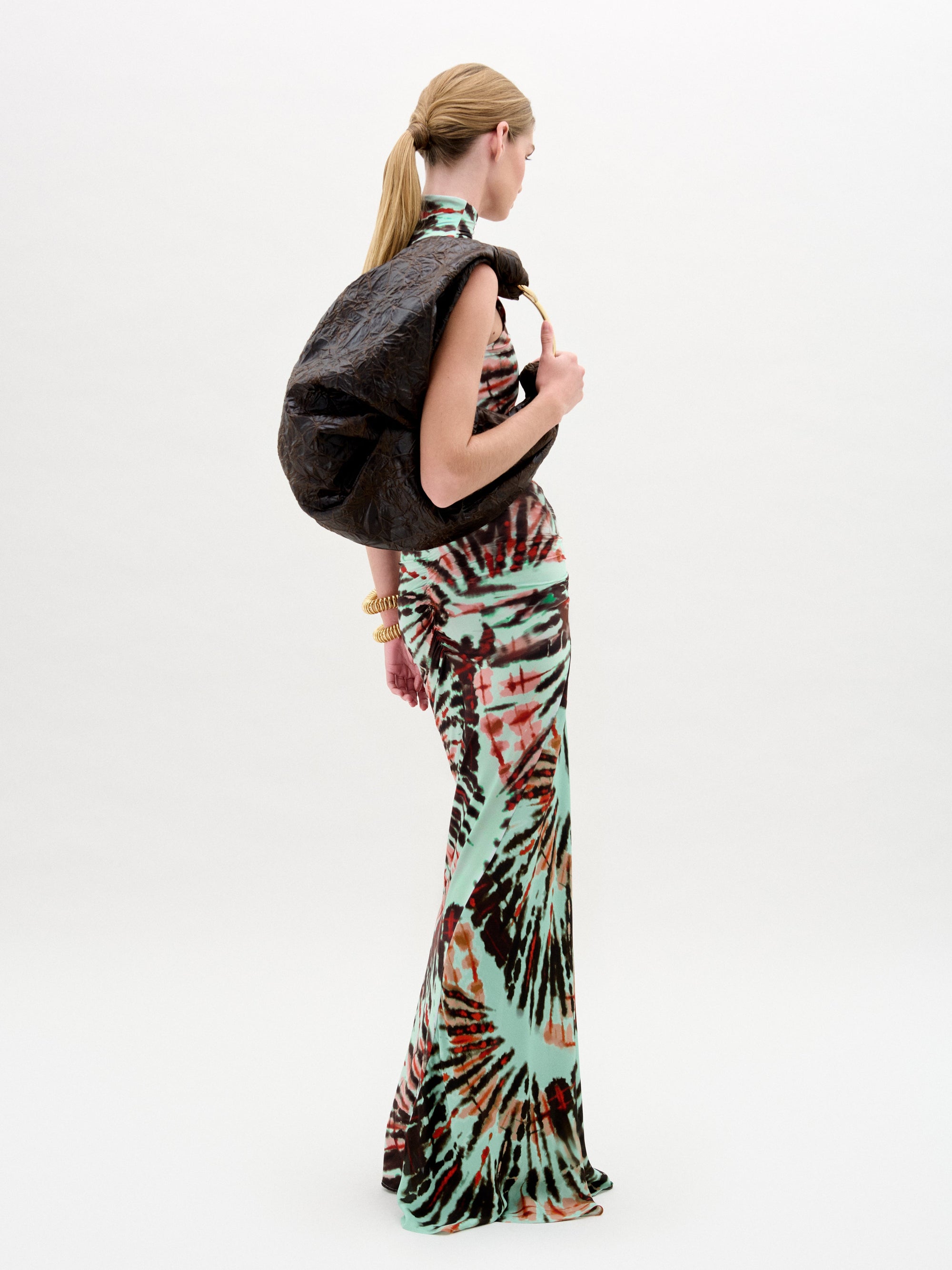 A person with long hair tied back is wearing the sleeveless, floor-length tie-dye Frances Dress by SVT DESIGNS. Made from Italian fabric, it drapes elegantly as a large, dark textured bag rests on their shoulder against a plain light-colored background.