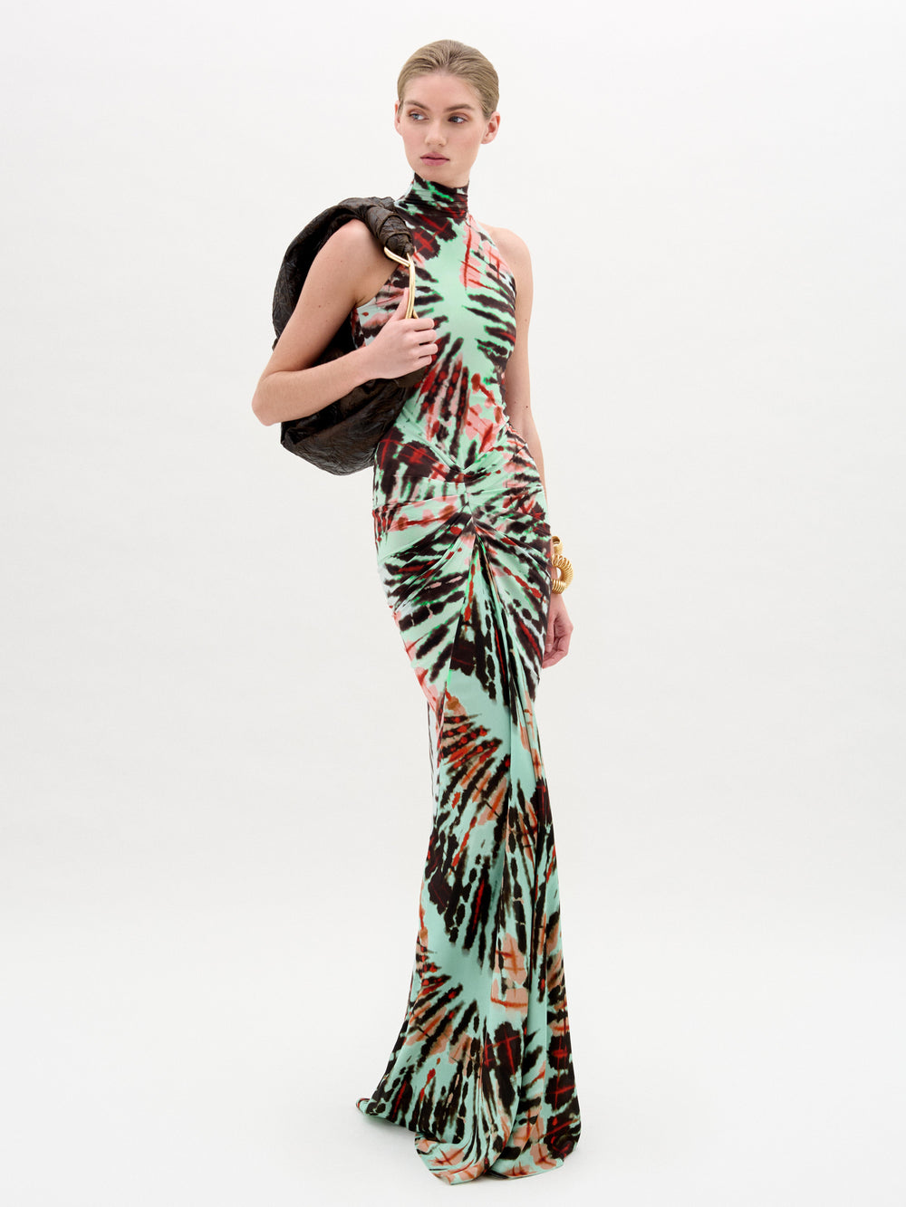 A woman poses against a plain backdrop in the SVT DESIGNS Frances Dress, a sleeveless, high-neck, slim maxi with green, black, and orange tie-dye tones, holding a textured dark jacket over her shoulder.