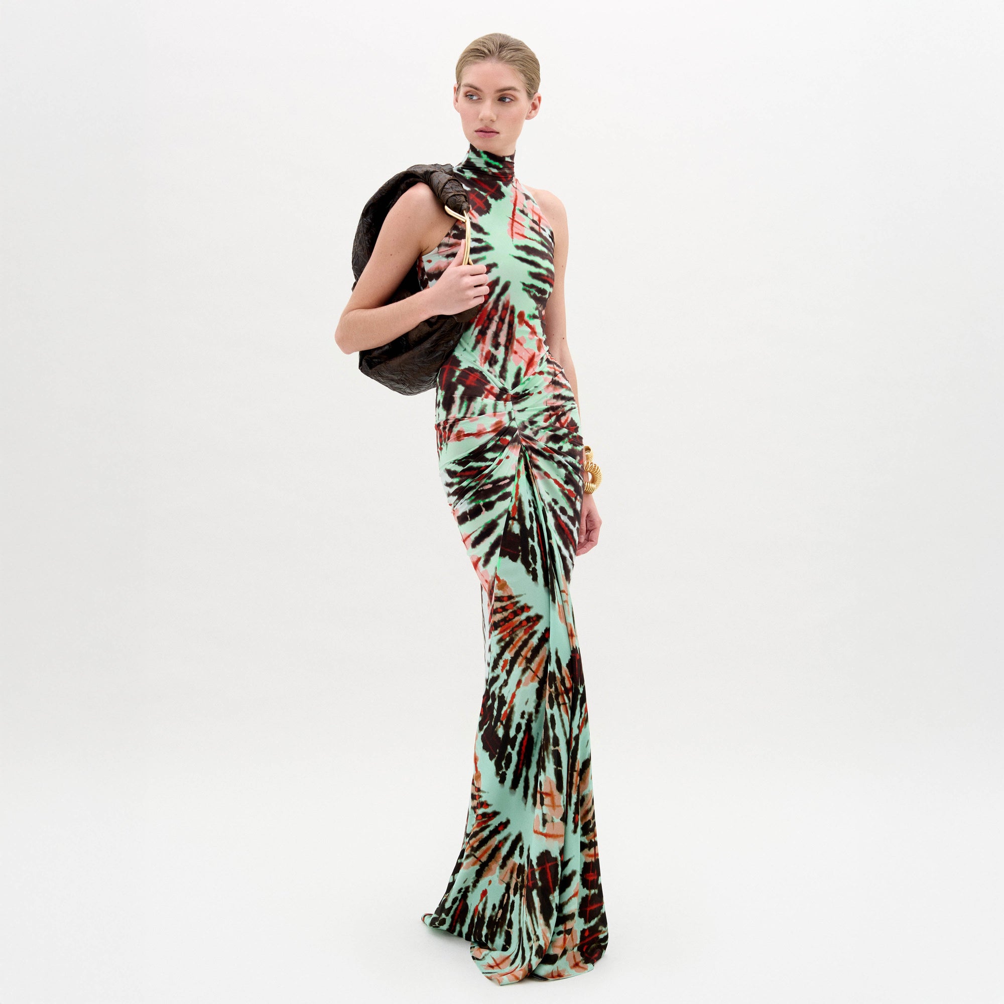 A woman wears the Frances Dress, a sleeveless, slim-fit maxi with a high neckline, holding a dark jacket over her shoulder against a plain white background.