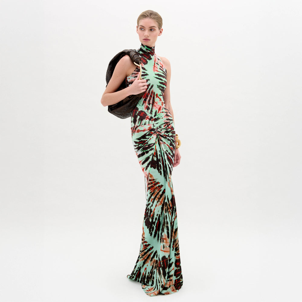 Against a plain white background, a person wears the SVT DESIGNS Frances Dress—a floor-length maxi with a multicolored tie-dye pattern and fitted bodice—holding a dark jacket over one shoulder and accessorizing with a gold bracelet.
