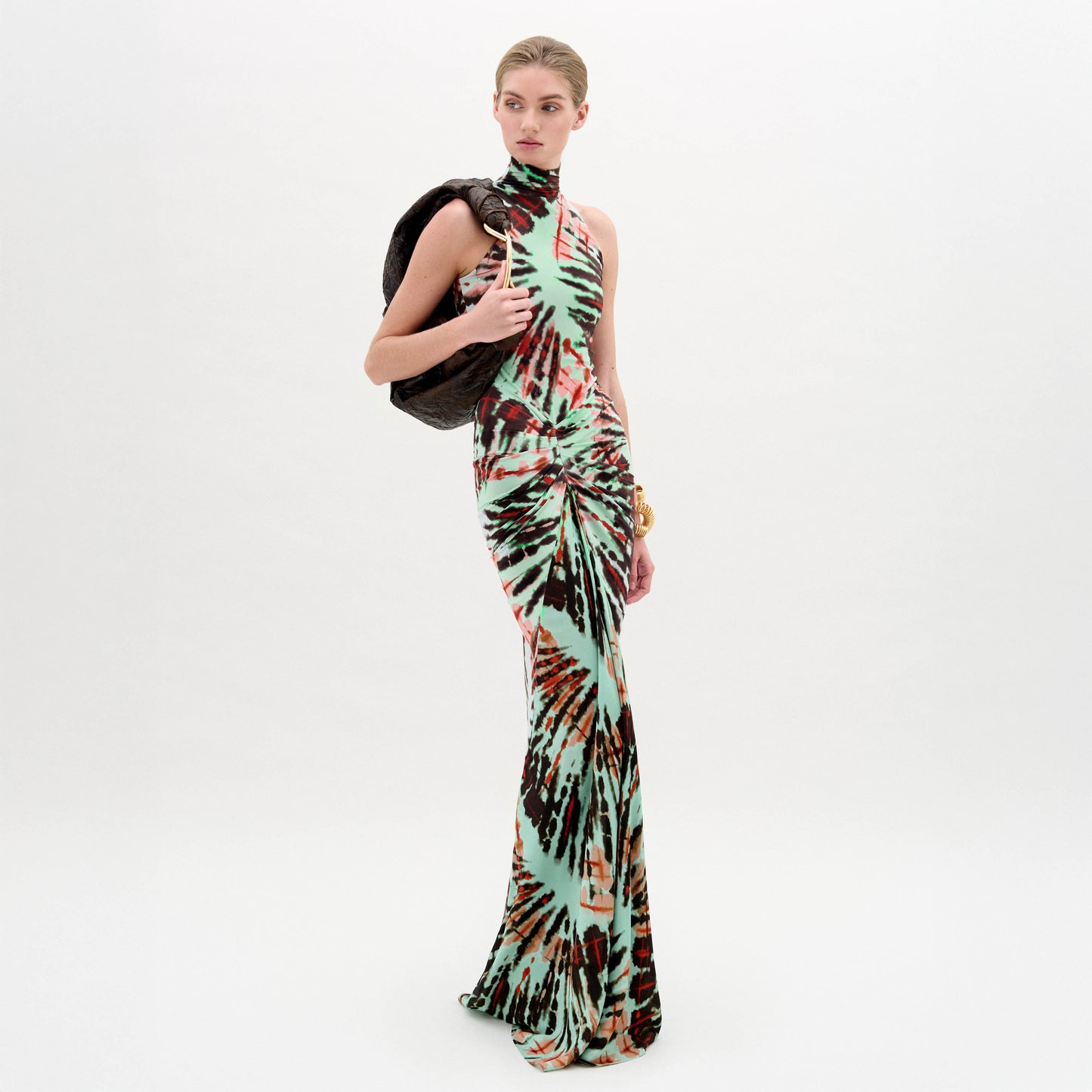 Against a plain white background, a person wears the SVT DESIGNS Frances Dress—a floor-length maxi with a multicolored tie-dye pattern and fitted bodice—holding a dark jacket over one shoulder and accessorizing with a gold bracelet.