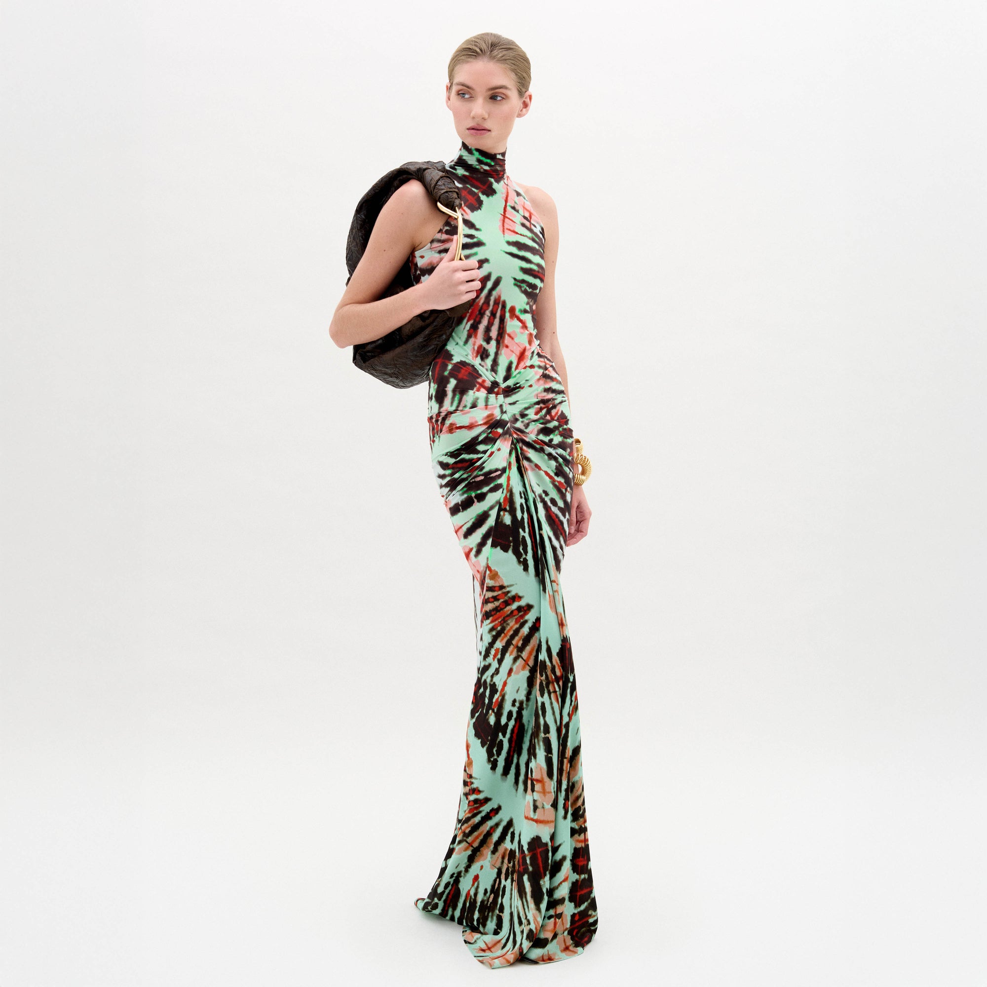 Against a plain white background, a person wears the SVT DESIGNS Frances Dress—a floor-length maxi with a multicolored tie-dye pattern and fitted bodice—holding a dark jacket over one shoulder and accessorizing with a gold bracelet.