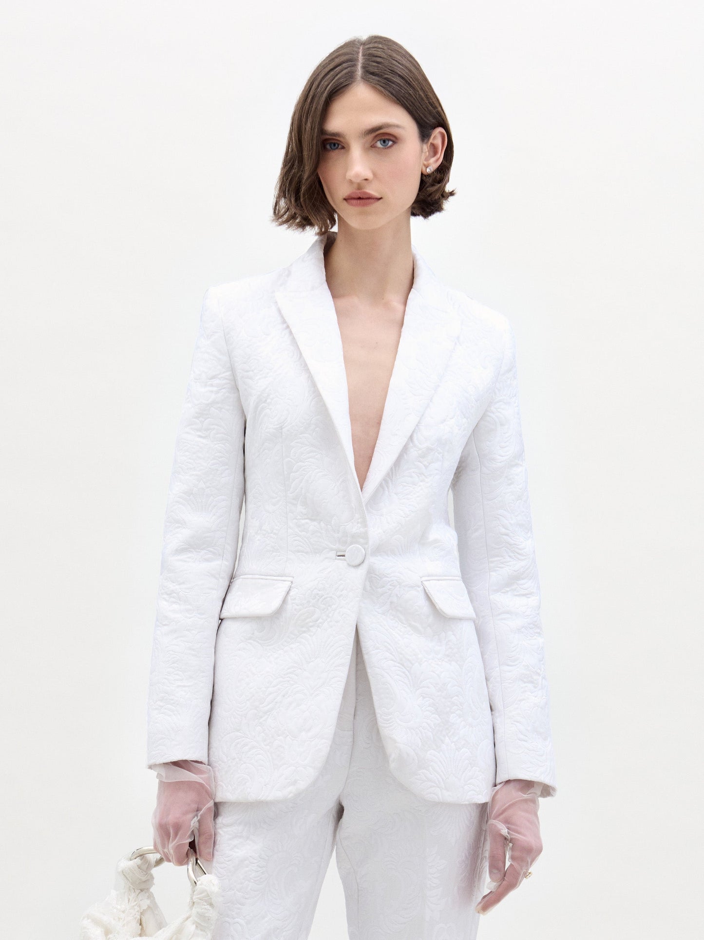 A woman with short brown hair wears the SVT DESIGNS Rebeca Jacket with matching pants, paired with sheer pale pink gloves and a small bag. She stands against a plain, light-colored background.
