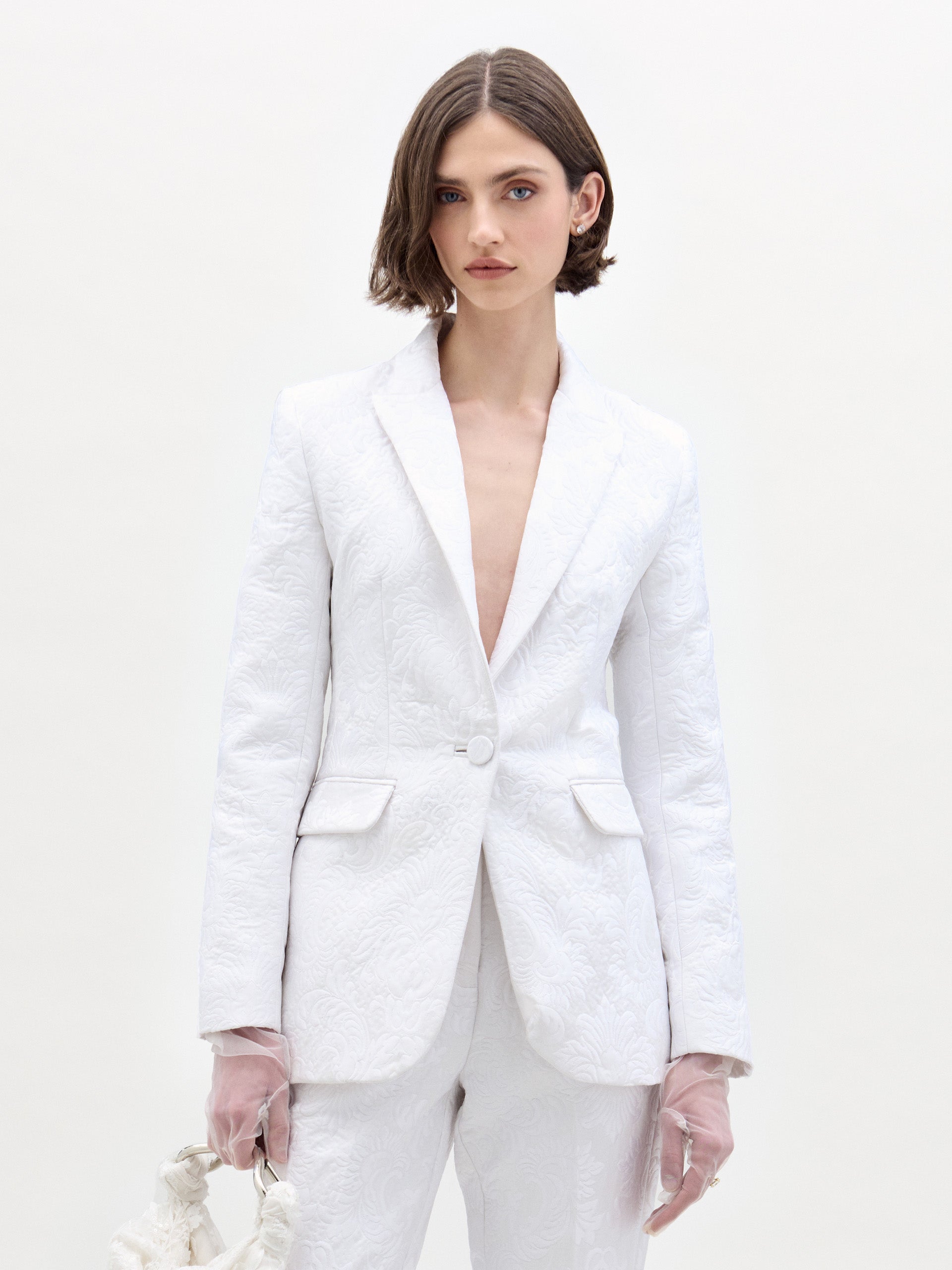 A woman with short brown hair wears the SVT DESIGNS Rebeca Jacket with matching pants, paired with sheer pale pink gloves and a small bag. She stands against a plain, light-colored background.