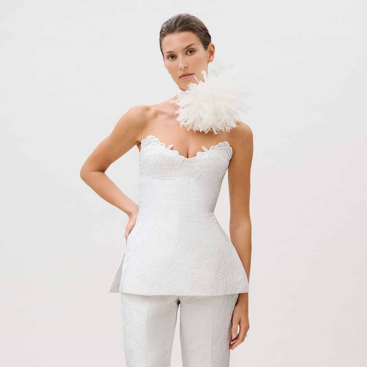 A woman wears the Filomena Top—a white, strapless, textured piece crafted from luxurious Italian fabric and finished with scalloped edges—paired with matching pants. She poses with a large white floral accessory on her shoulder against a plain light background, showcasing this ready-to-wear ensemble that exudes sophistication and timeless elegance.