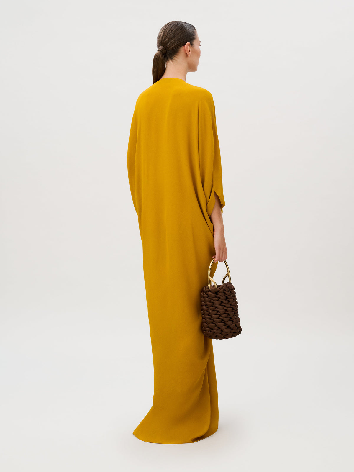 A person wearing a mustard-yellow Fiore Tunic with long, wide sleeves, holding a dark brown, chunky woven bag with a gold metal handle.