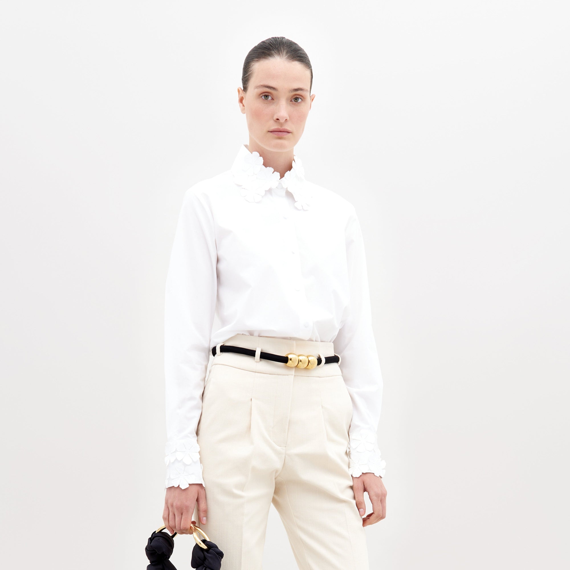 A person stands against a plain white background wearing the SVT DESIGNS Flower Blouse (100% cotton, floral appliqué cuffs), cream trousers, a black and gold belt, and holding a small black item in their left hand.