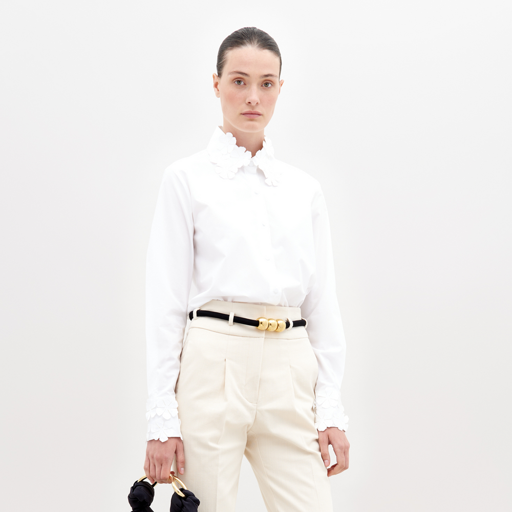 A woman with dark hair pulled back models the Flower Blouse—100% cotton with floral details on the collar and cuffs—paired with cream pants and a black belt with a gold buckle, standing against a plain white background.