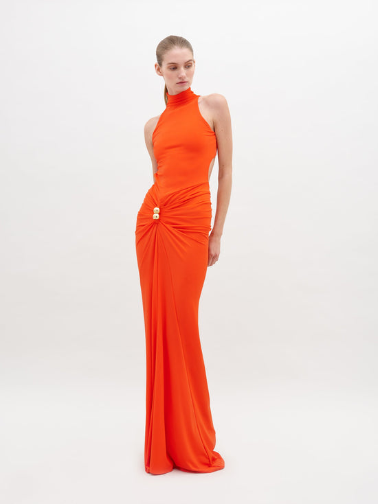 A woman in a long, sleeveless, high-neck orange Frances Dress Tangerine stands against a plain white background. The dress is gathered at the waist with a decorative clasp. This stylish piece is available for pre-order now and will ship by November 15th, 2024.