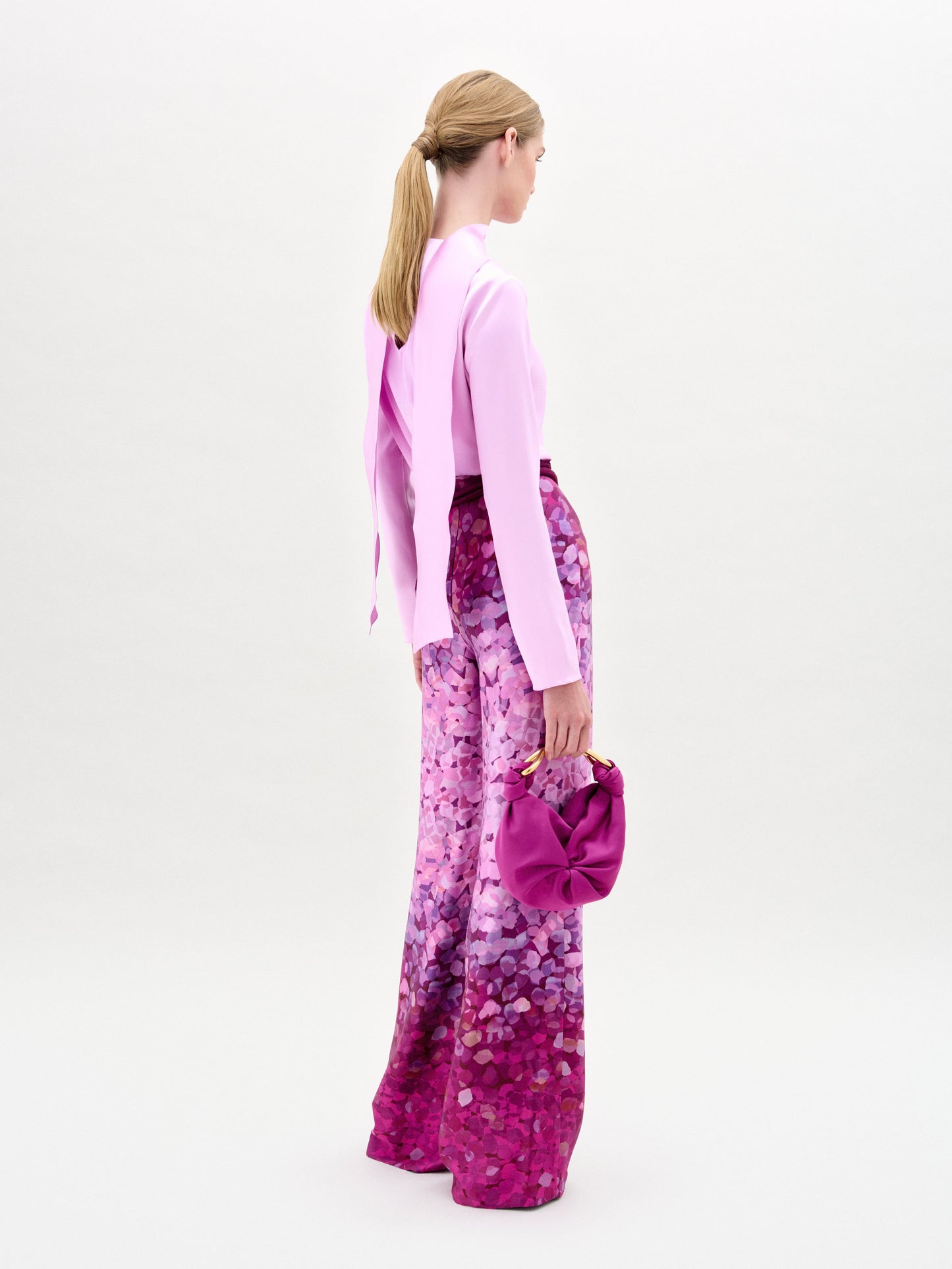 A woman stands against a plain background wearing a light pink long-sleeve top and the Como Pant—high-waisted, wide-leg pants crafted from Italian fabric—while holding a matching small bag.