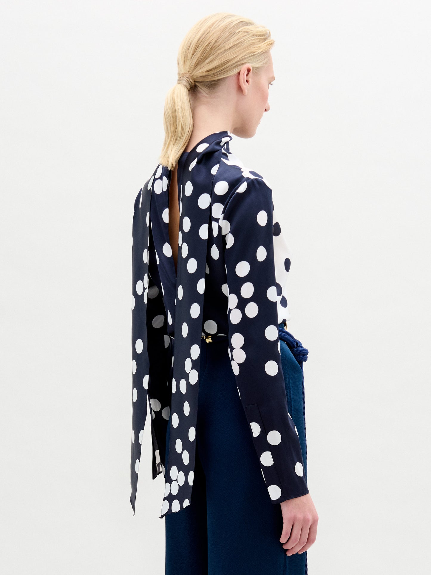 A person with blond hair tied back is elegantly dressed in a SVT DESIGNS Gaby Blouse, made from Italian fabric in navy blue with large white polka dots and an open back design. It's paired with dark blue high-waisted pants against a plain, light-colored background.