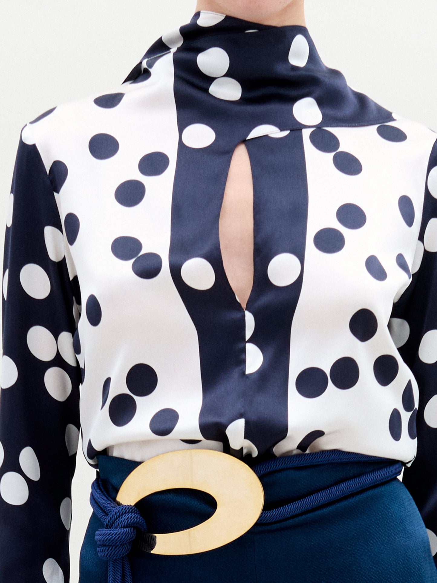 A person is wearing the Gaby Blouse by SVT DESIGNS, featuring a navy blue and white polka dot silk fabric with an elegant keyhole neckline and high collar. They also sport a dark blue belt with a large oval wooden buckle, highlighting the charm of Italian craftsmanship.