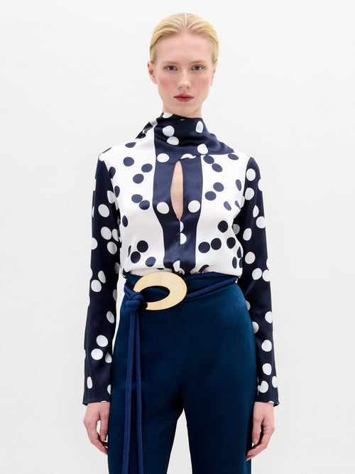 A person is wearing the SVT DESIGNS Gaby Blouse, a high-neck silk top with large black and white polka dots and a keyhole neckline. They’ve paired it with dark blue pants featuring a large circular beige buckle and a matching fabric belt. The background is plain white.