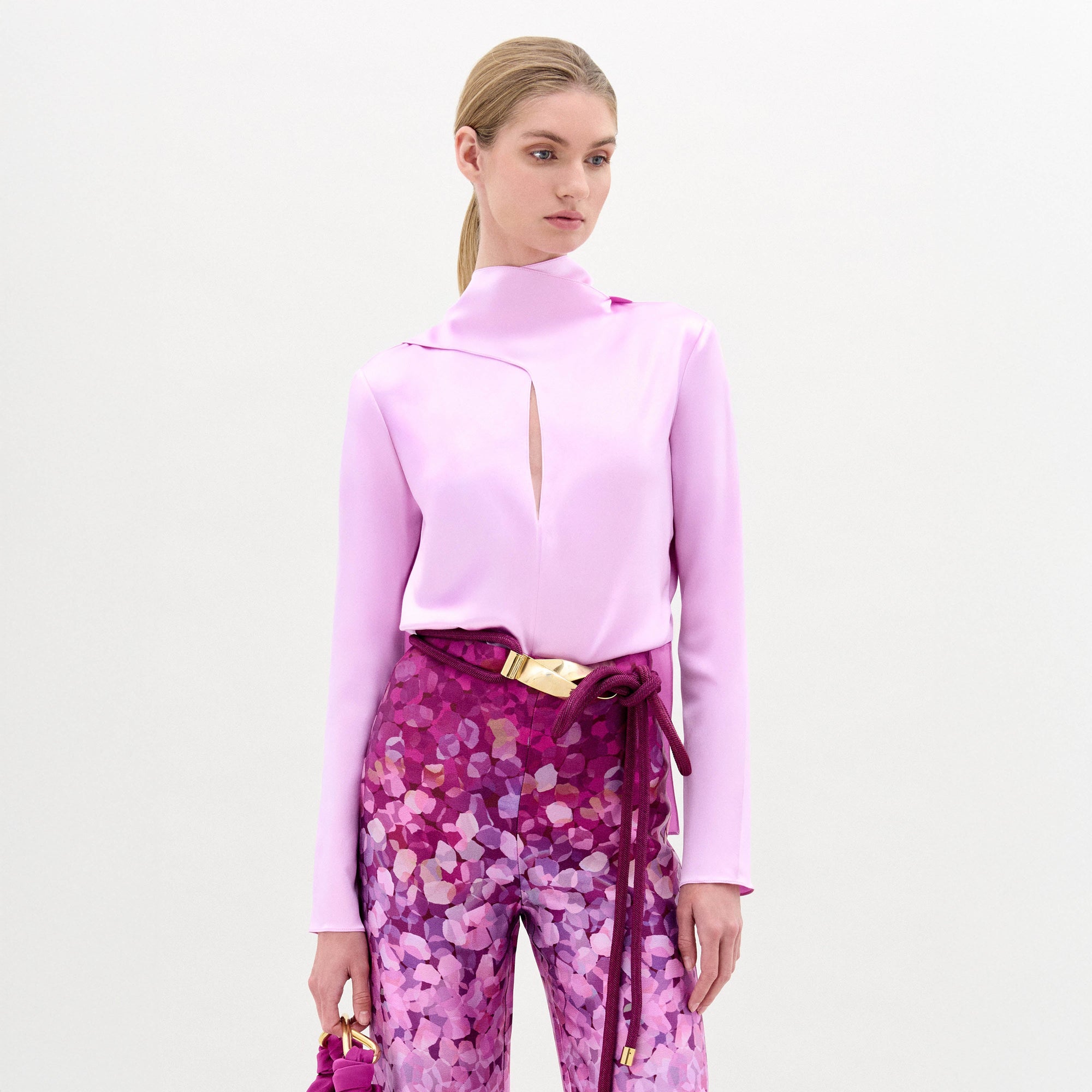 A woman poses against a plain background in the Gaby Blouse, a light pink silk long-sleeve top with a front slit, paired with high-waisted pink and purple patterned pants, a wide belt, and a small matching handbag.