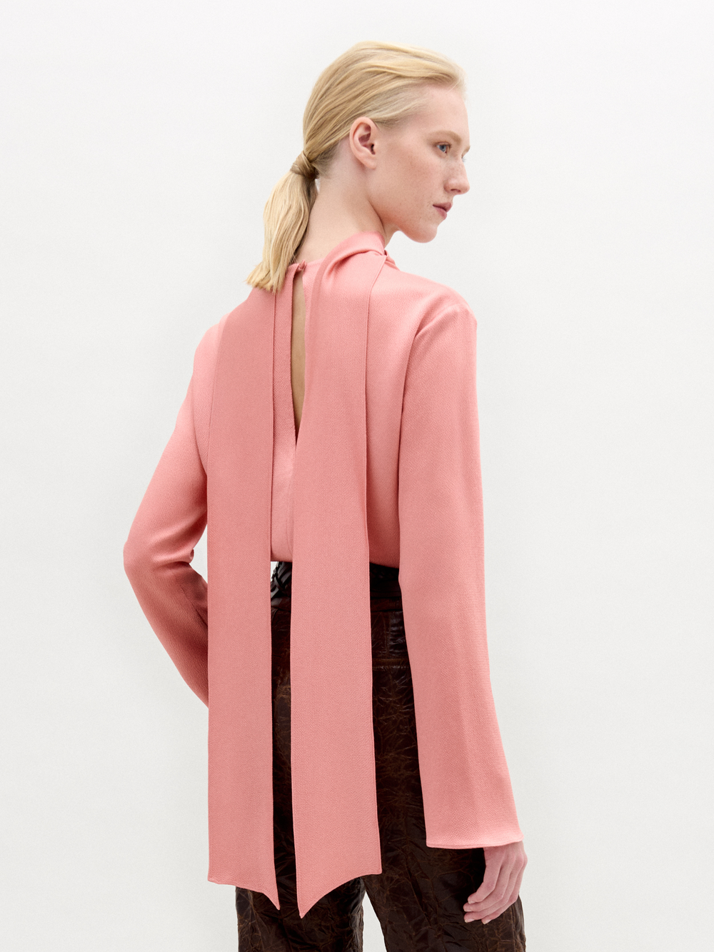 A woman stands against a plain background wearing the Gaby Blouse, a pink high-neck top with a front slit, paired with brown textured pants and a wide belt featuring a large gold oval buckle.