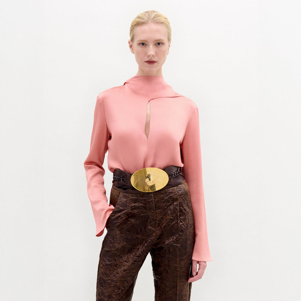 A woman stands against a plain background wearing the Gaby Blouse, a pink high-neck top with a front slit, paired with brown textured pants and a wide belt featuring a large gold oval buckle.