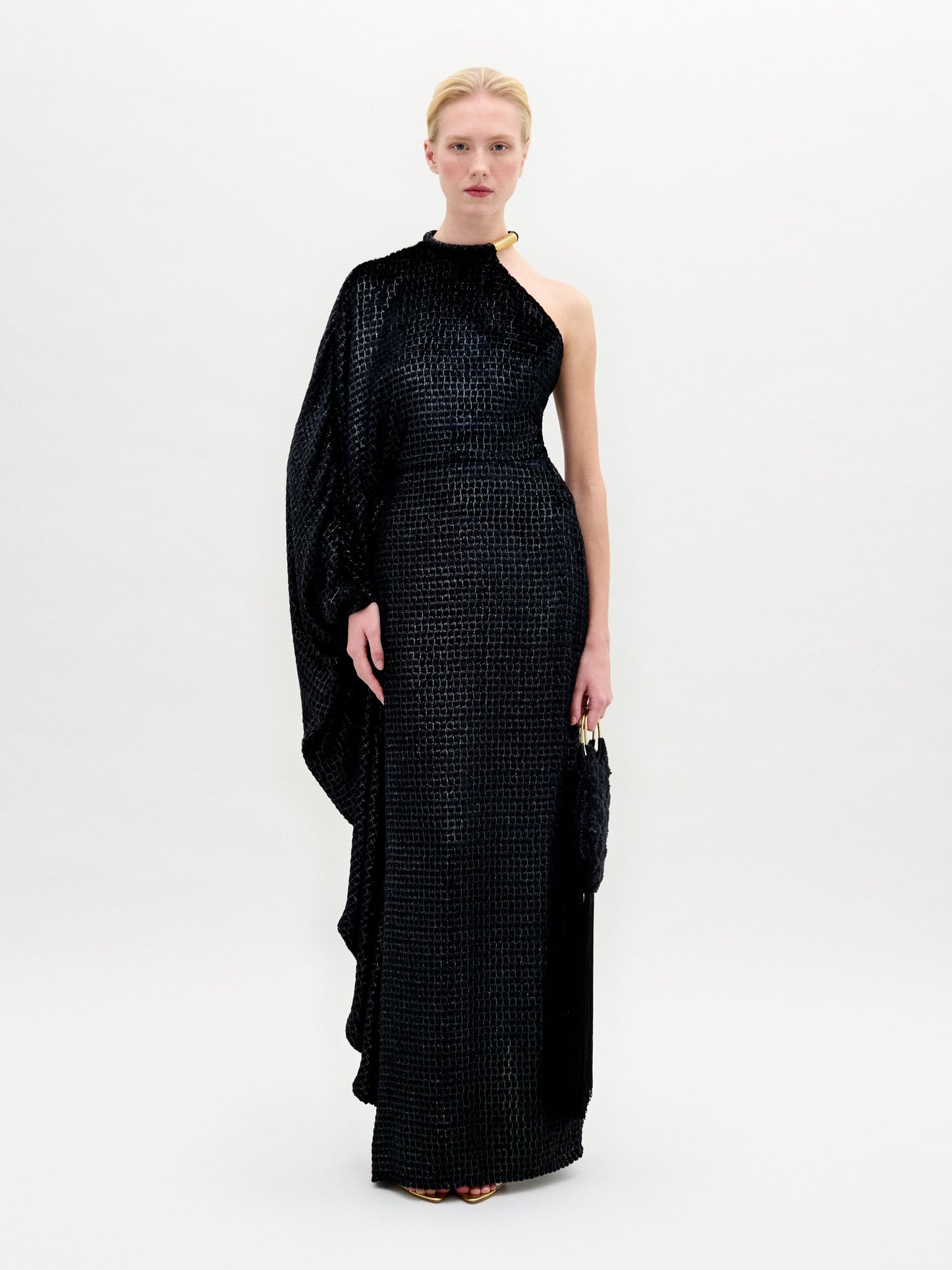 A person stands against a plain white background wearing the SVT DESIGNS Gael Dress—a floor-length maxi with an asymmetrical neckline in textured black Italian fabric—holding a small black clutch and with hair pulled back.