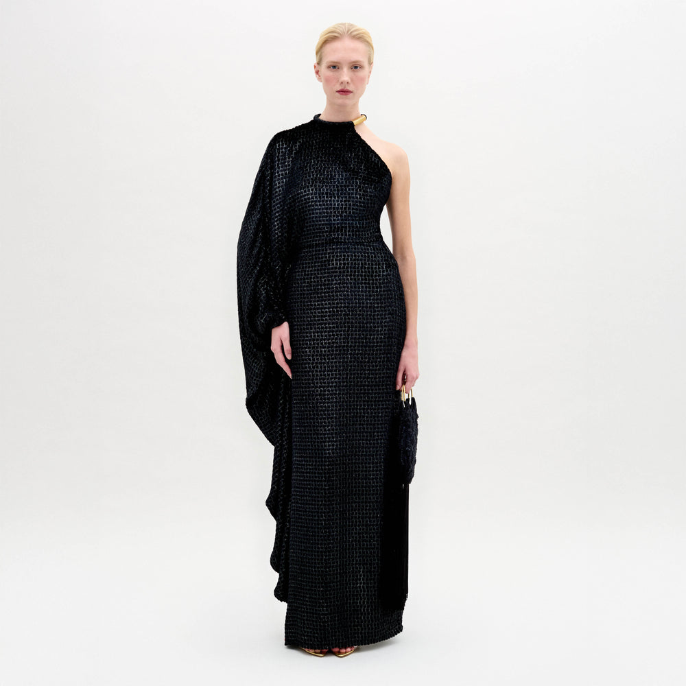 A person stands against a white background wearing the Gael Dress by SVT DESIGNS—a long, one-shoulder black maxi in textured Italian fabric featuring an asymmetrical neckline with a draped sleeve—holding a small black clutch.