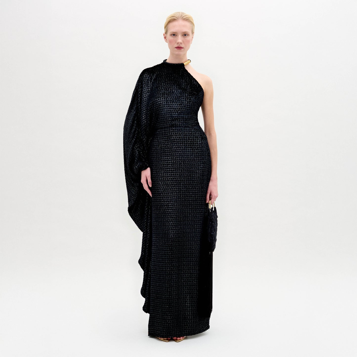 A person stands against a white background wearing the Gael Dress by SVT DESIGNS—a long, one-shoulder black maxi in textured Italian fabric featuring an asymmetrical neckline with a draped sleeve—holding a small black clutch.