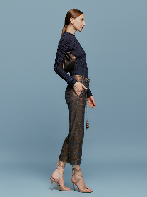 A woman stands against a blue background, wearing a textured navy long-sleeve top, Garmet Pant Metallic Ocher pants, and strappy nude heels, holding a brown clutch bag. She embodies the essence of effortless elegance on August 19th.