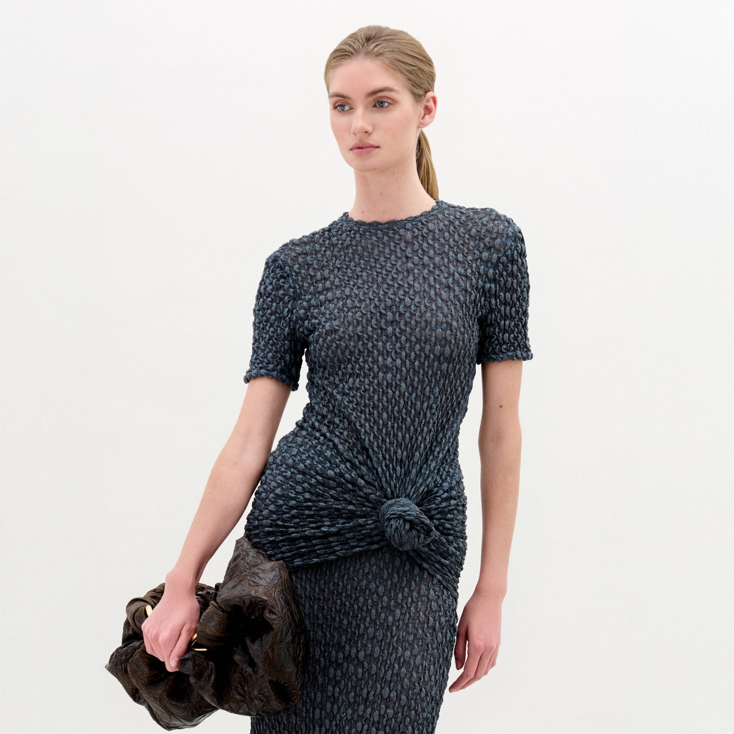 A woman with blonde hair in a low ponytail wears the Gen Tshirt, a textured, fitted, dark short-sleeve top with knot detail, and holds a large dark ruched bag against a plain white background.