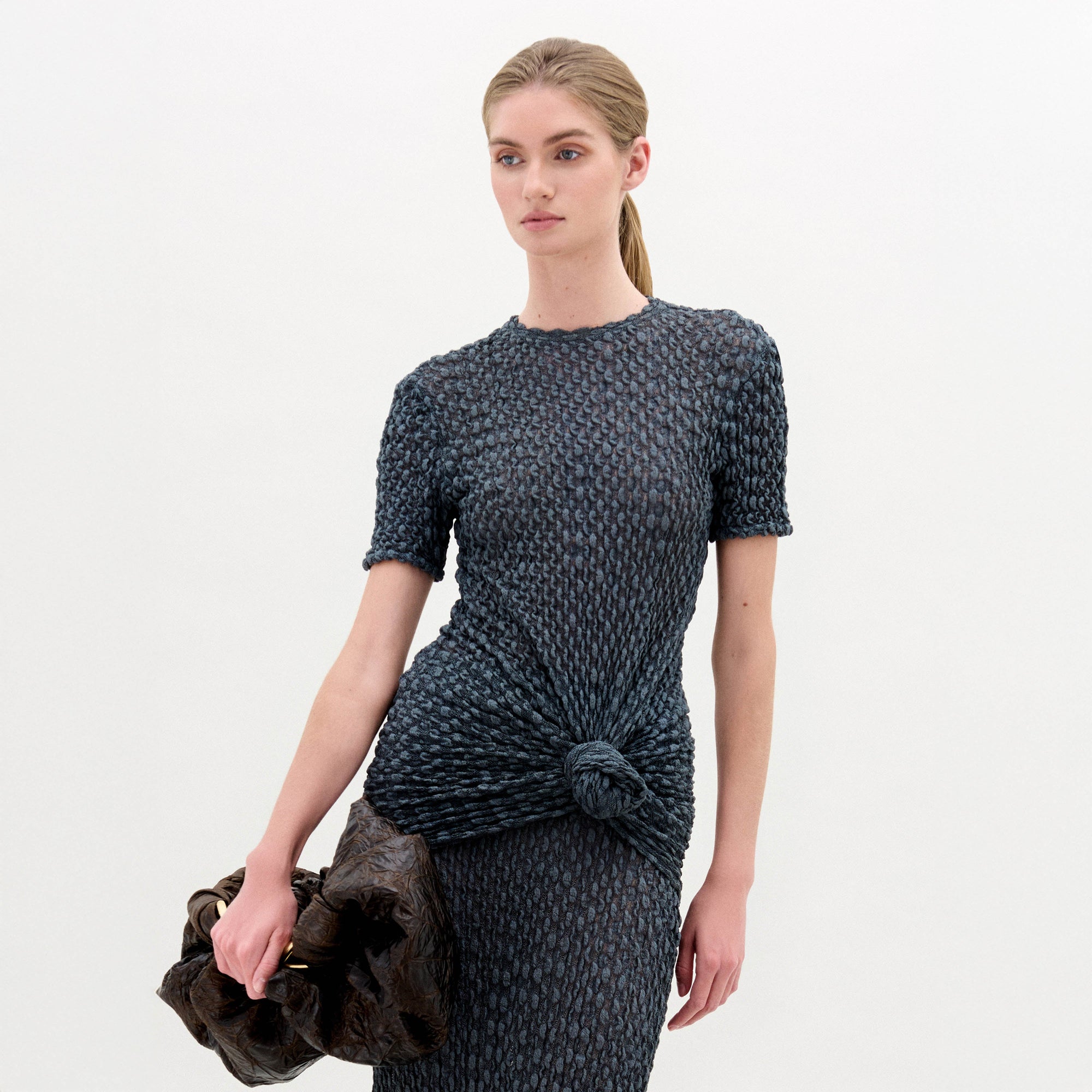 A woman with blonde hair in a low ponytail wears the Gen Tshirt, a textured, fitted, dark short-sleeve top with knot detail, and holds a large dark ruched bag against a plain white background.