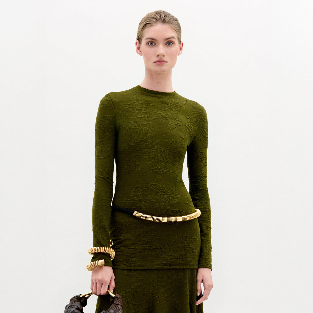 A woman with blonde hair tied back models the SVT DESIGNS Gen Tshirt, styled with a black and gold belt and a sculptural bag featuring gold details, all set against a plain white background.