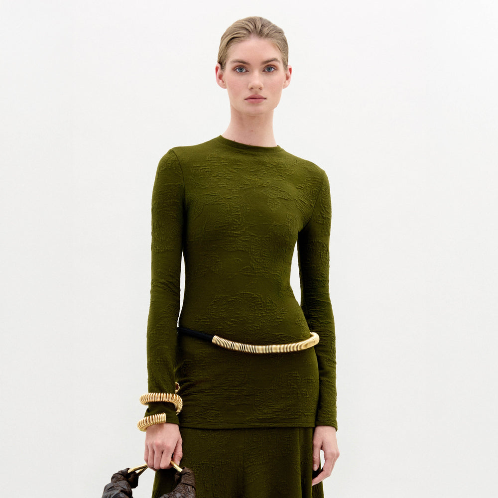 A woman stands against a plain white background wearing the Gen Tshirt, a slim fit, textured olive green long-sleeve dress with gold accessories and holding a brown bag.