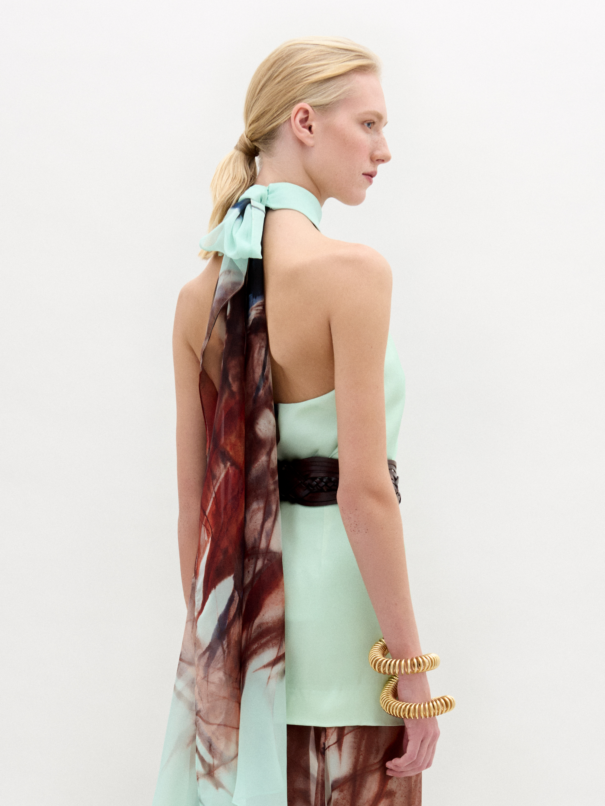 A woman stands against a plain white background wearing the Geovanna Blouse—a sleeveless, high-neck halter top featuring a brown and light blue abstract pattern—paired with a wide belt and matching skirt, made in Colombia.