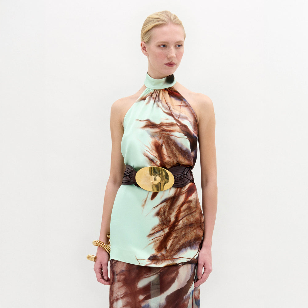 A woman stands against a plain white background wearing the Geovanna Blouse—a sleeveless, high-neck halter top featuring a brown and light blue abstract pattern—paired with a wide belt and matching skirt, made in Colombia.