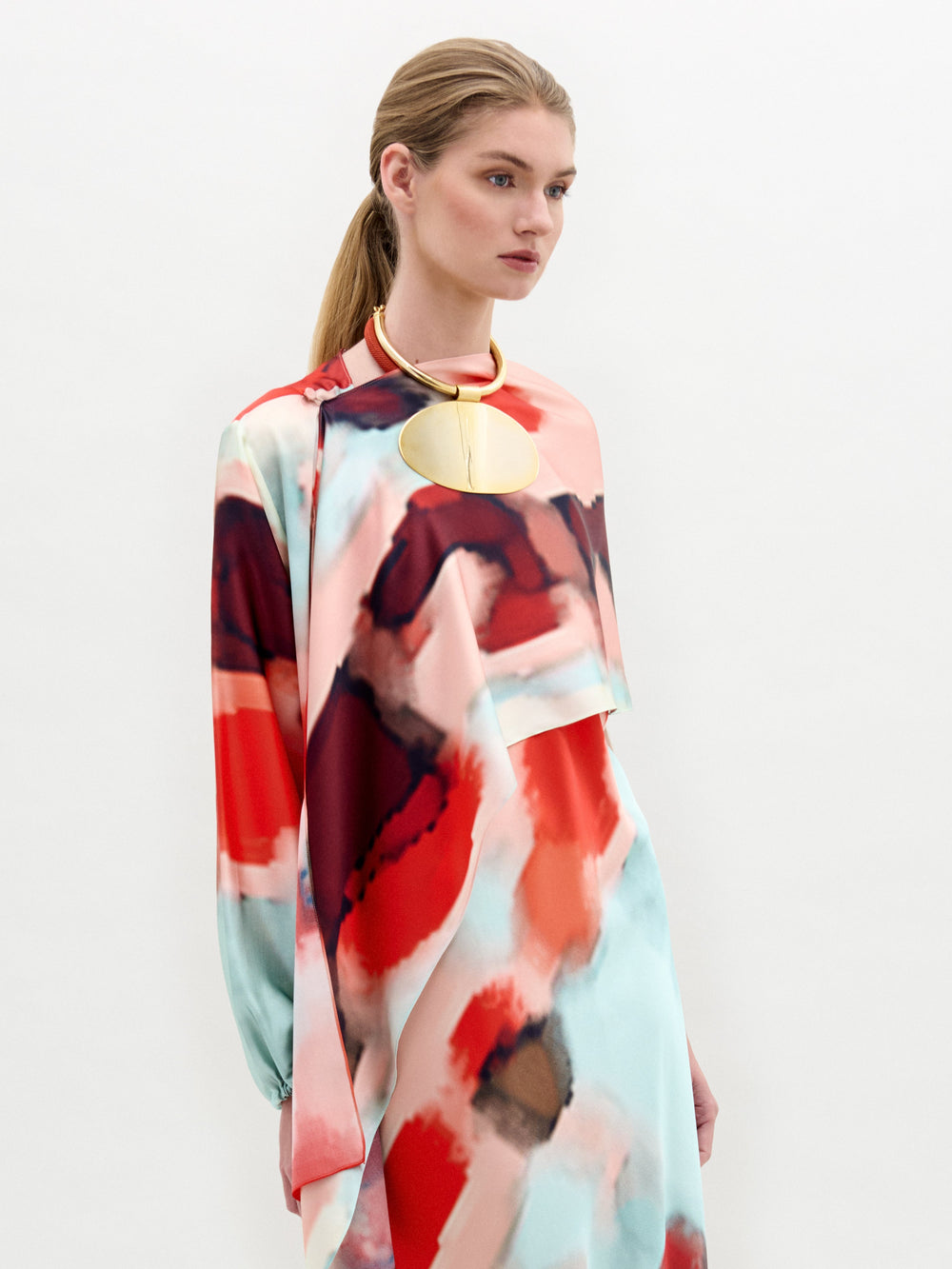 A person with long hair tied back stands against a plain white background wearing the Gery Shrug by SVT DESIGNS. This long-sleeve dress is made from exquisite Italian fabric, featuring a vibrant red, pink, and blue abstract pattern with a large circular gold accessory at the neckline.