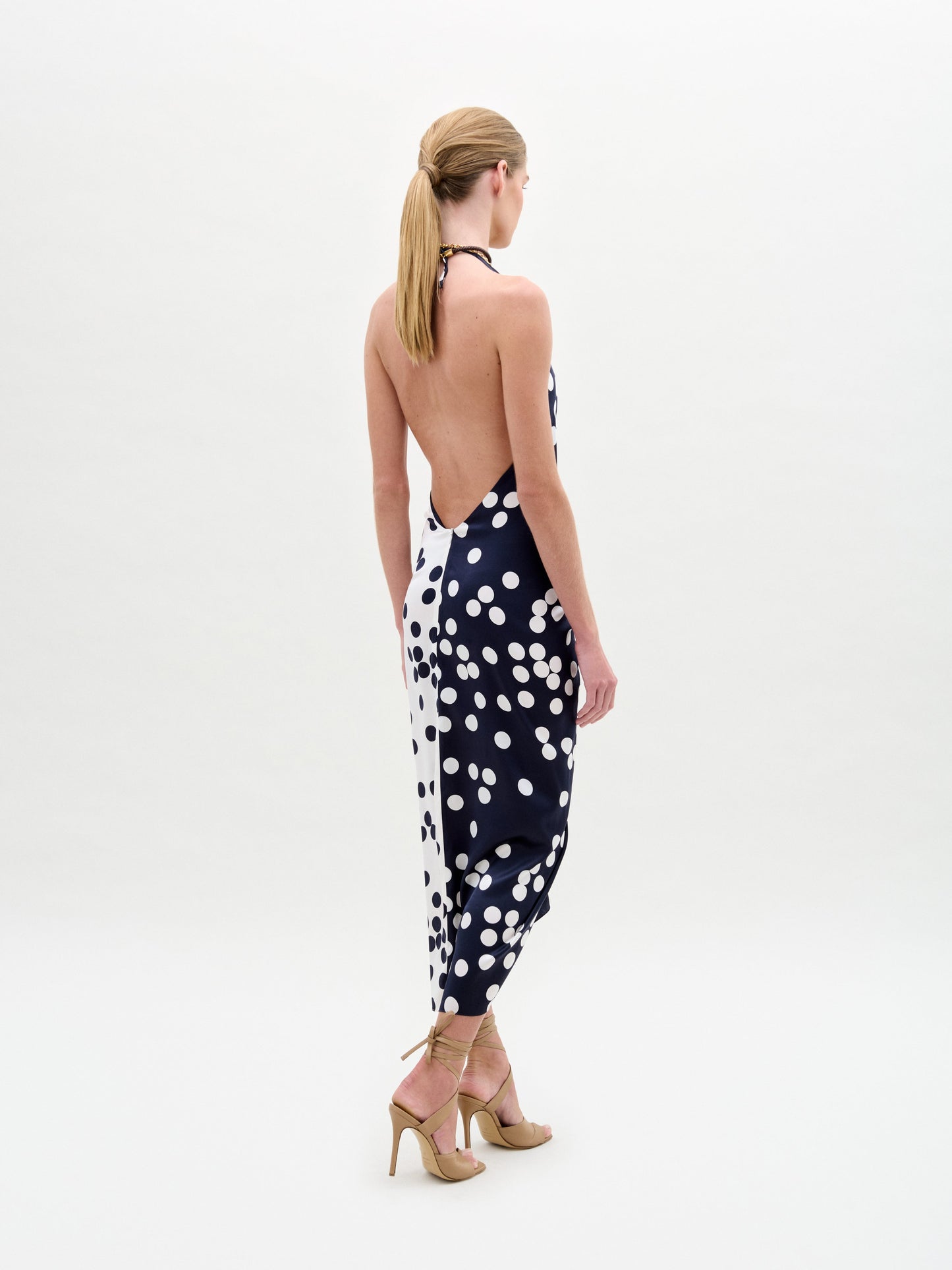 Facing away, a person with long hair in a ponytail wears the Guadalupe Dress by SVT DESIGNS. The silk midi dress features a draped silhouette, deep V halter neckline, and black and white polka dots. She pairs it elegantly with tan high-heeled sandals against a plain white background.