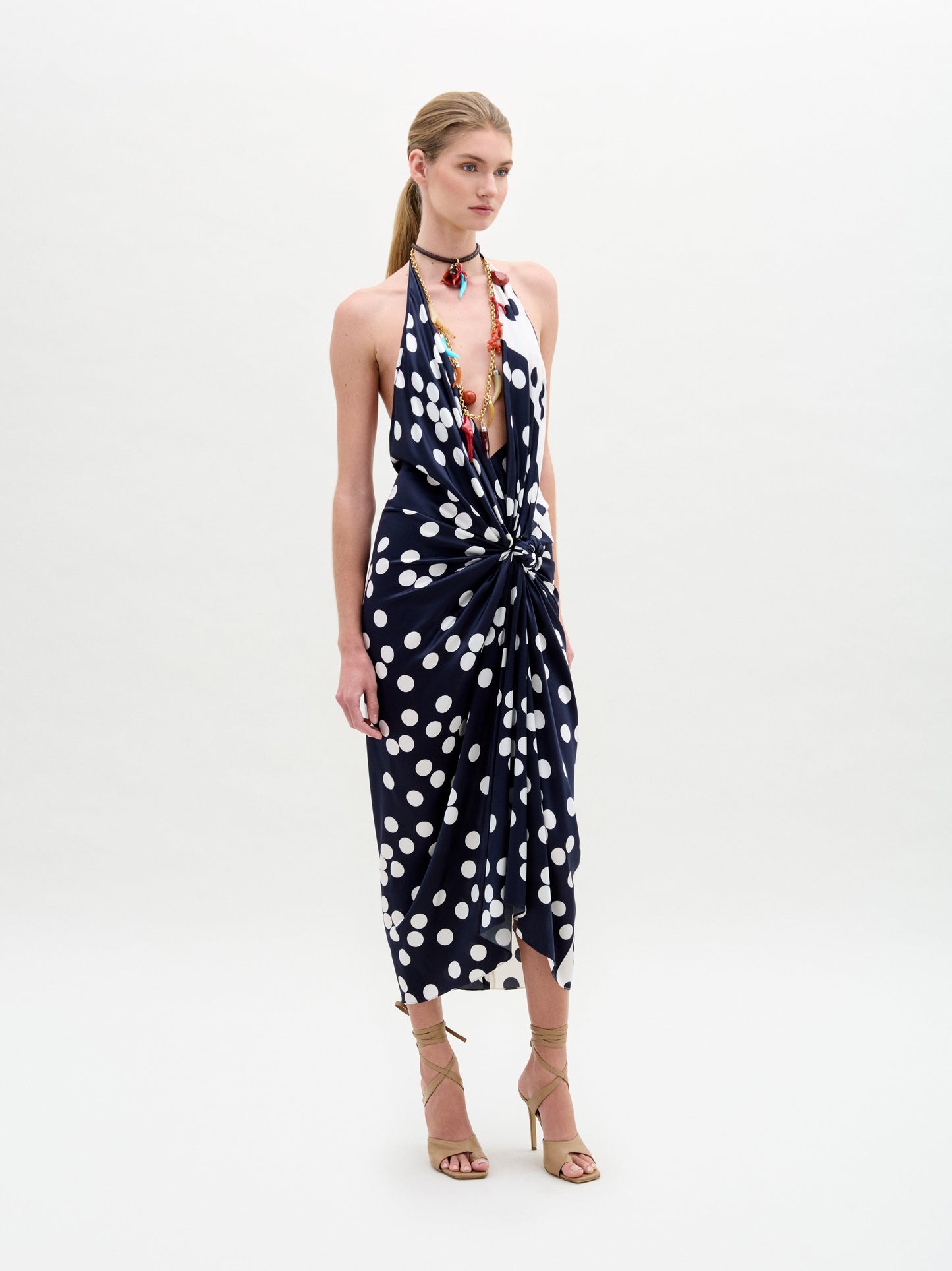 A woman stands against a plain background wearing the SVT DESIGNS Guadalupe Dress, a sleeveless silk navy blue midi dress with white polka dots. The dress has a deep V halter neckline and is knotted at the waist for a draped look. She adds a choker necklace, strappy high-heeled sandals, and ties her hair back.