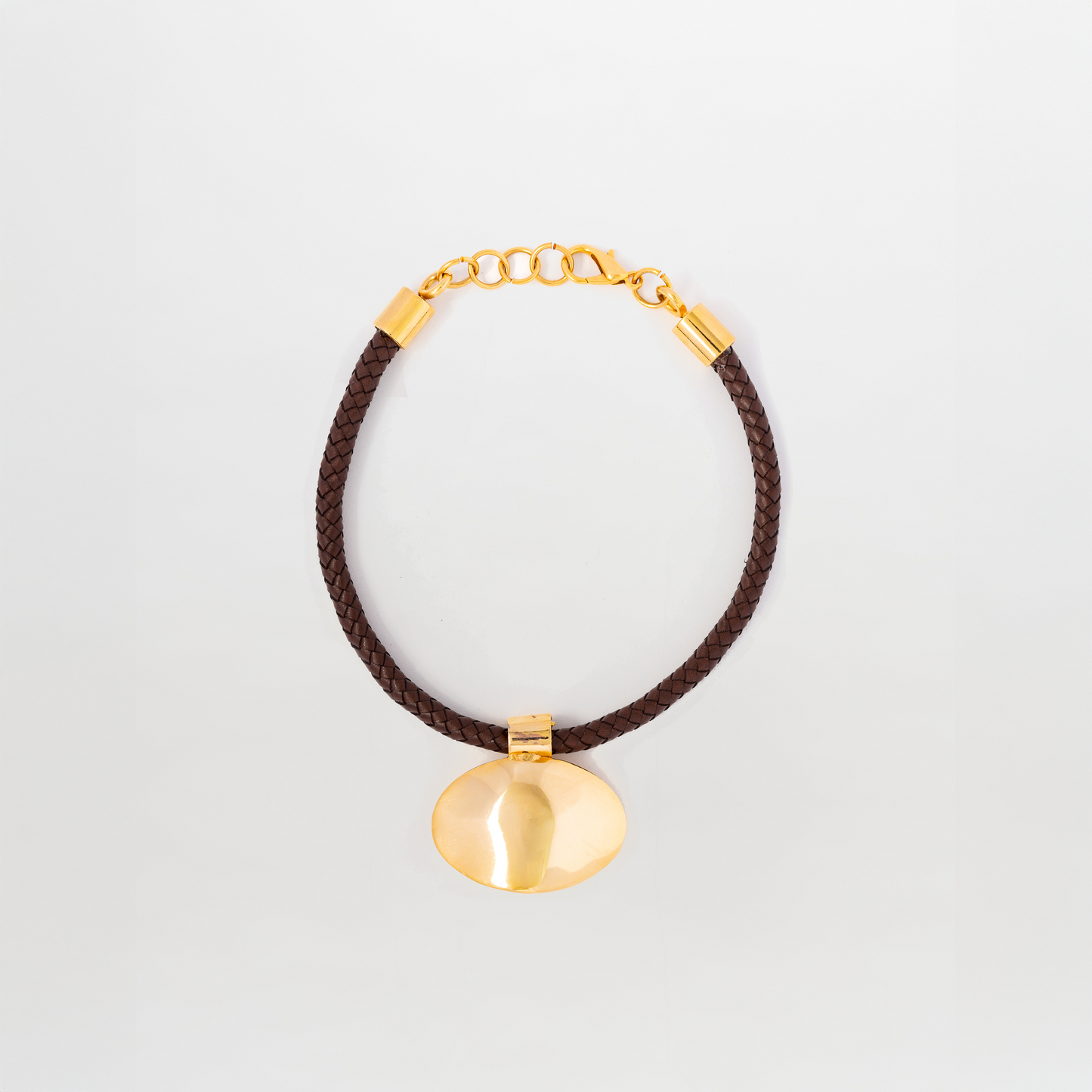 The Gimena Necklace by SVT DESIGNS features a gold pendant with a smooth, rounded shape on a dark brown braided cord with gold accents and an adjustable clasp, shown against a plain white background.