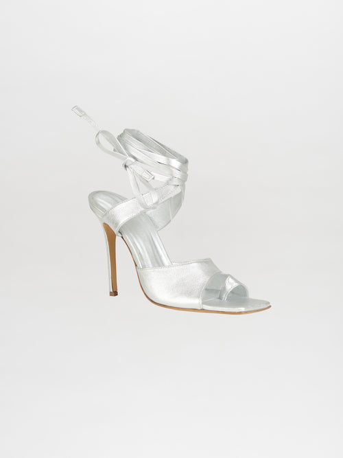 The Golda Heels Silver by Silvia Tcherassi features metallic silver straps, an open toe, and elegant lace-up ankle details. The sandals have a thin stiletto heel and are made from quality leather, showcased on a white background.