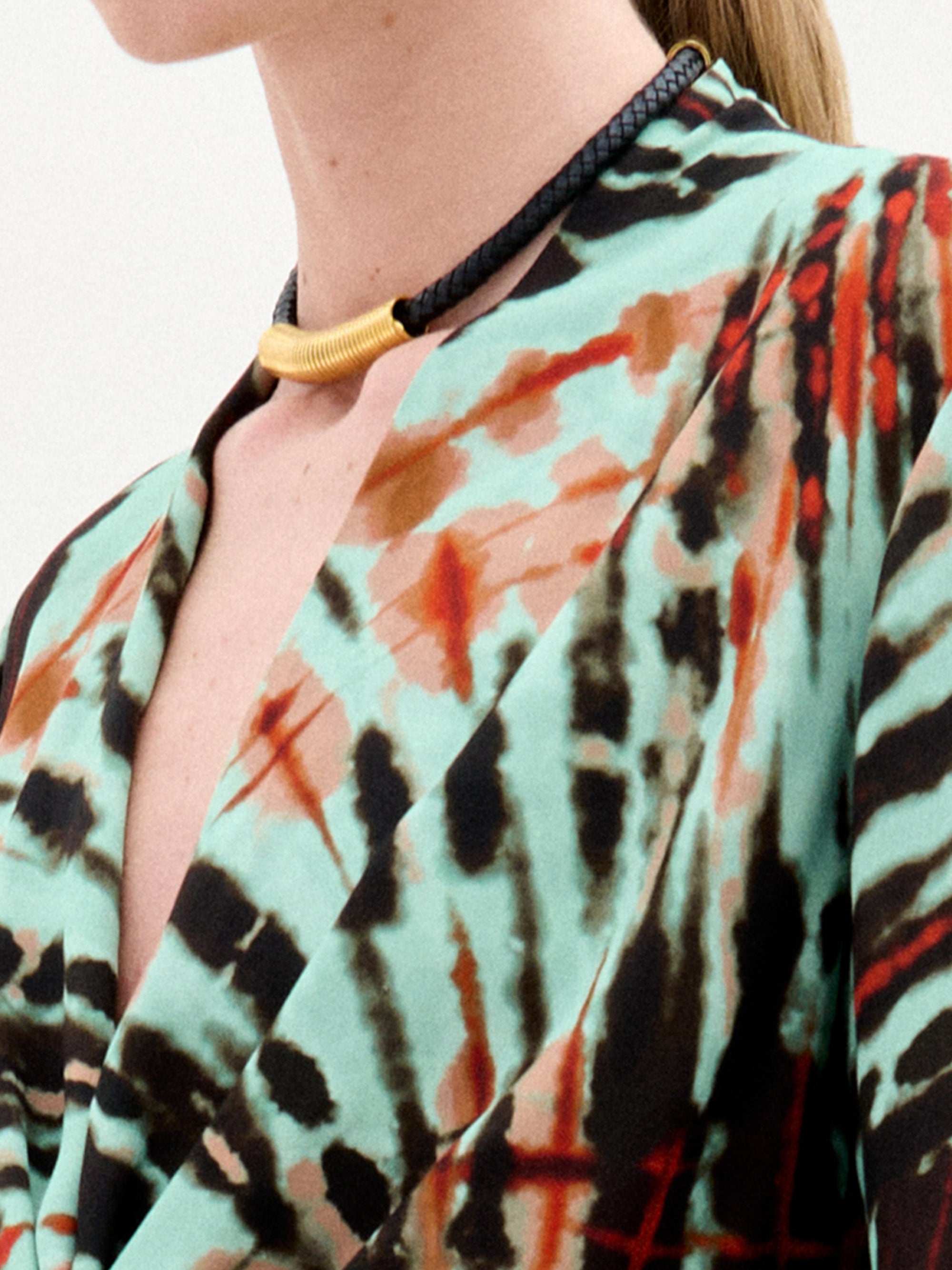 A person wears the Hadria Necklace by SVT DESIGNS—a short leather necklace with a gold curved accent—paired with a colorful turquoise, orange, and black garment. Only their neck, shoulder, and upper chest are visible. Made in Colombia.