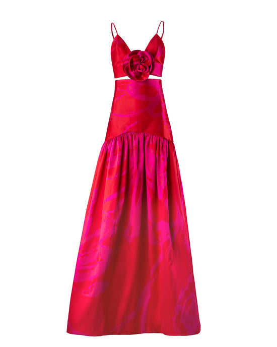 A person is standing wearing the Helen Set Dress Rouge Fuchsia Flower, a red, sleeveless gown featuring a floral detail on the bodice and a long skirt. The background is plain white. This stunning piece will be available for pre-order soon, with an expected ship-by date in early 2025.