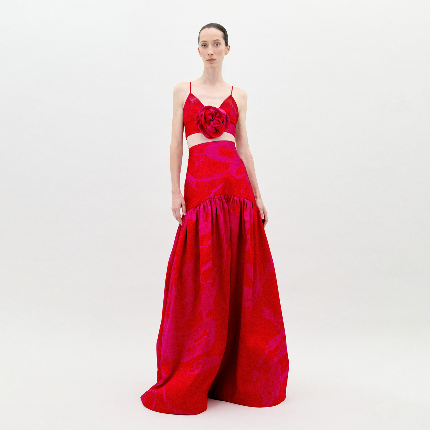 A person is standing wearing the Helen Set Dress Rouge Fuchsia Flower, a red, sleeveless gown featuring a floral detail on the bodice and a long skirt. The background is plain white. This stunning piece will be available for pre-order soon, with an expected ship-by date in early 2025.