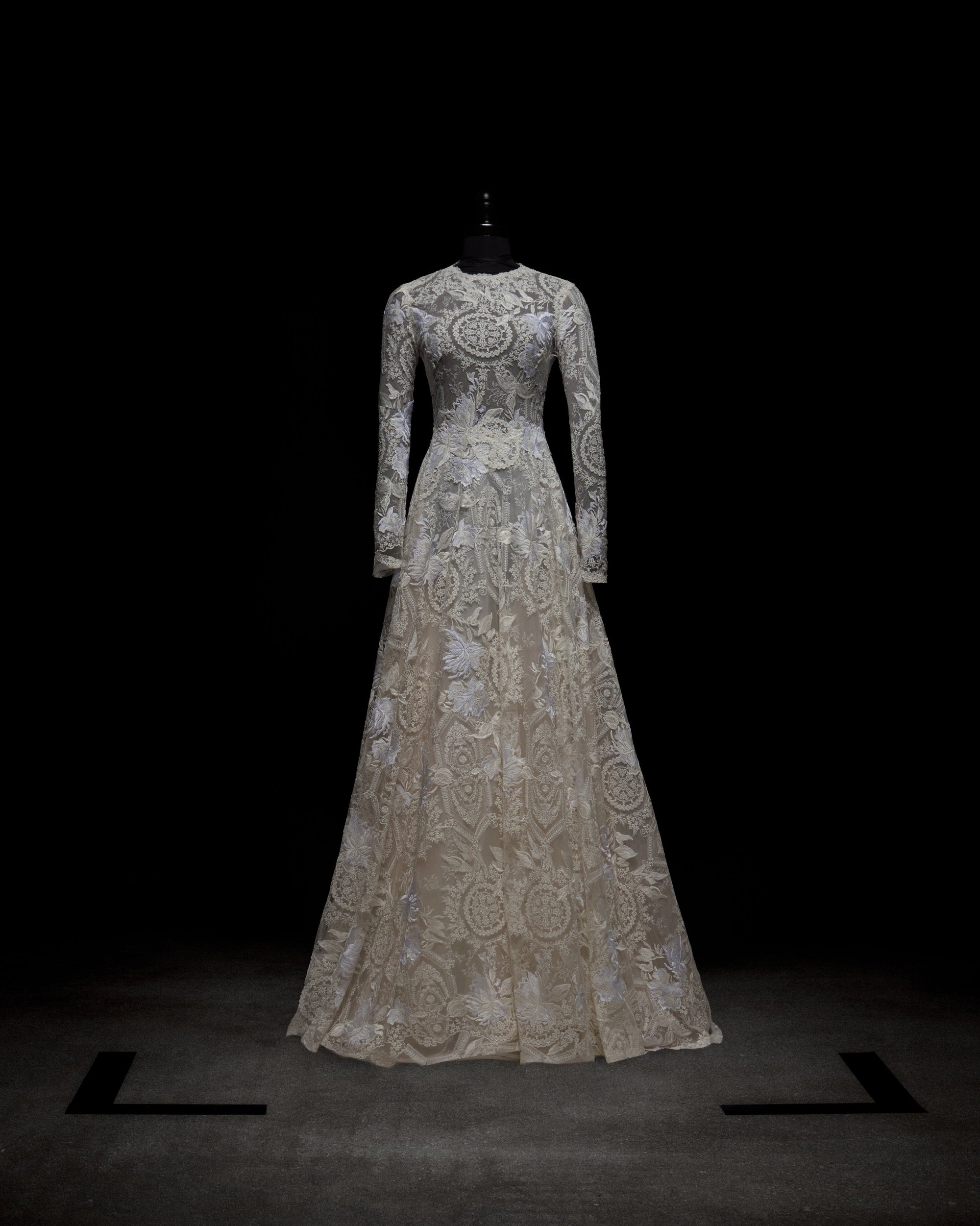 The Hermione Dress by SVT DESIGNS is showcased on a black mannequin against a dark background. It features intricate lace patterns on its long sleeves and fitted bodice that beautifully lead to a flowing, full-length skirt.