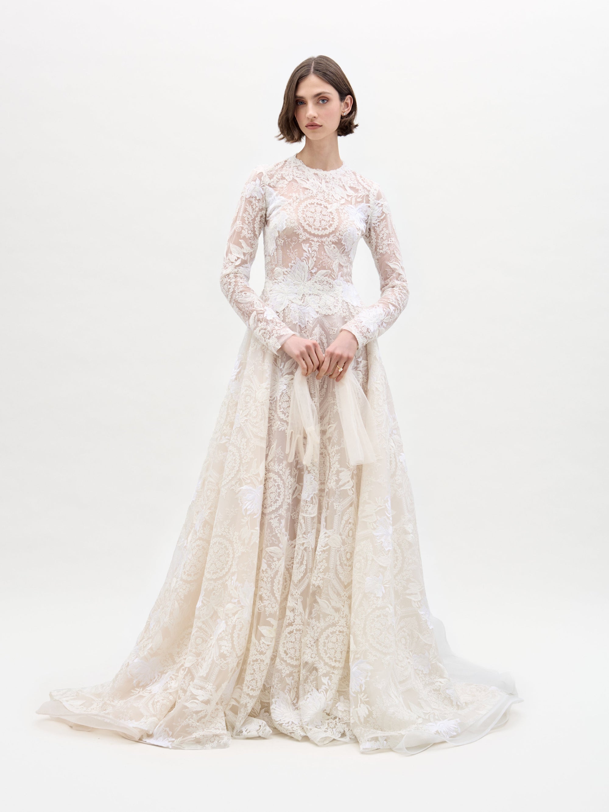 Against a plain white backdrop, an individual in the SVT DESIGNS Hermione Dress, a long cream-colored lace gown with intricate patterns and long sleeves, stands. The person has shoulder-length hair and gently holds the gown's lower part.