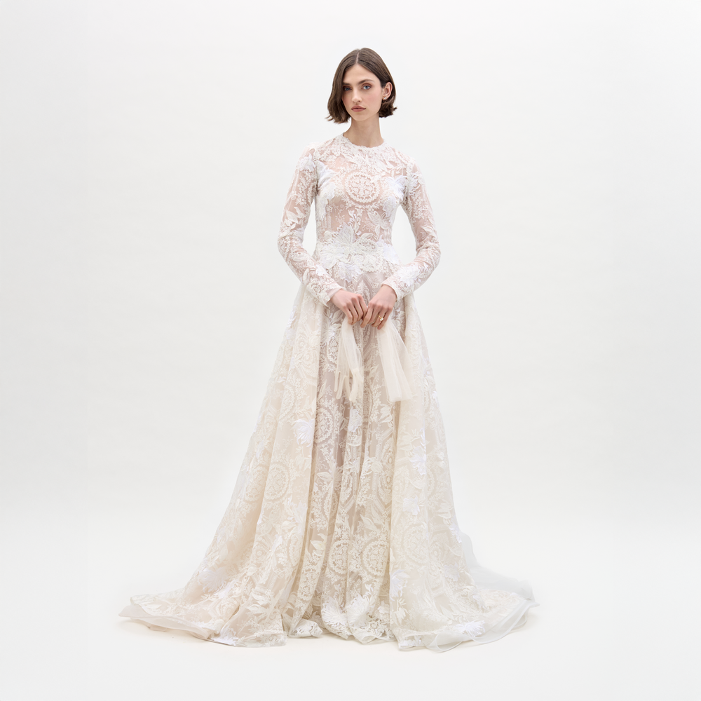 A woman wears the Hermione Dress by SVT DESIGNS, a floor-length, long-sleeved lace wedding gown with intricate floral embroidery. She holds a light fabric while posing against a plain white background.