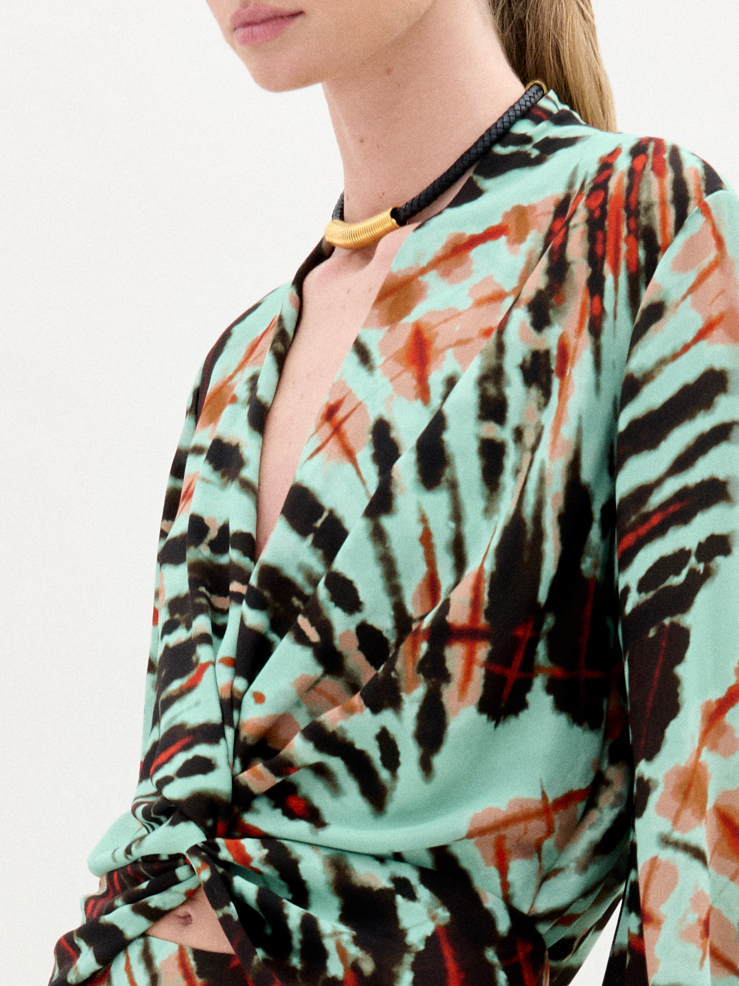 A woman stands against a plain white background wearing the Honey Blouse, a long-sleeved top featuring green, black, and orange abstract patterns with a chic front twist detail.