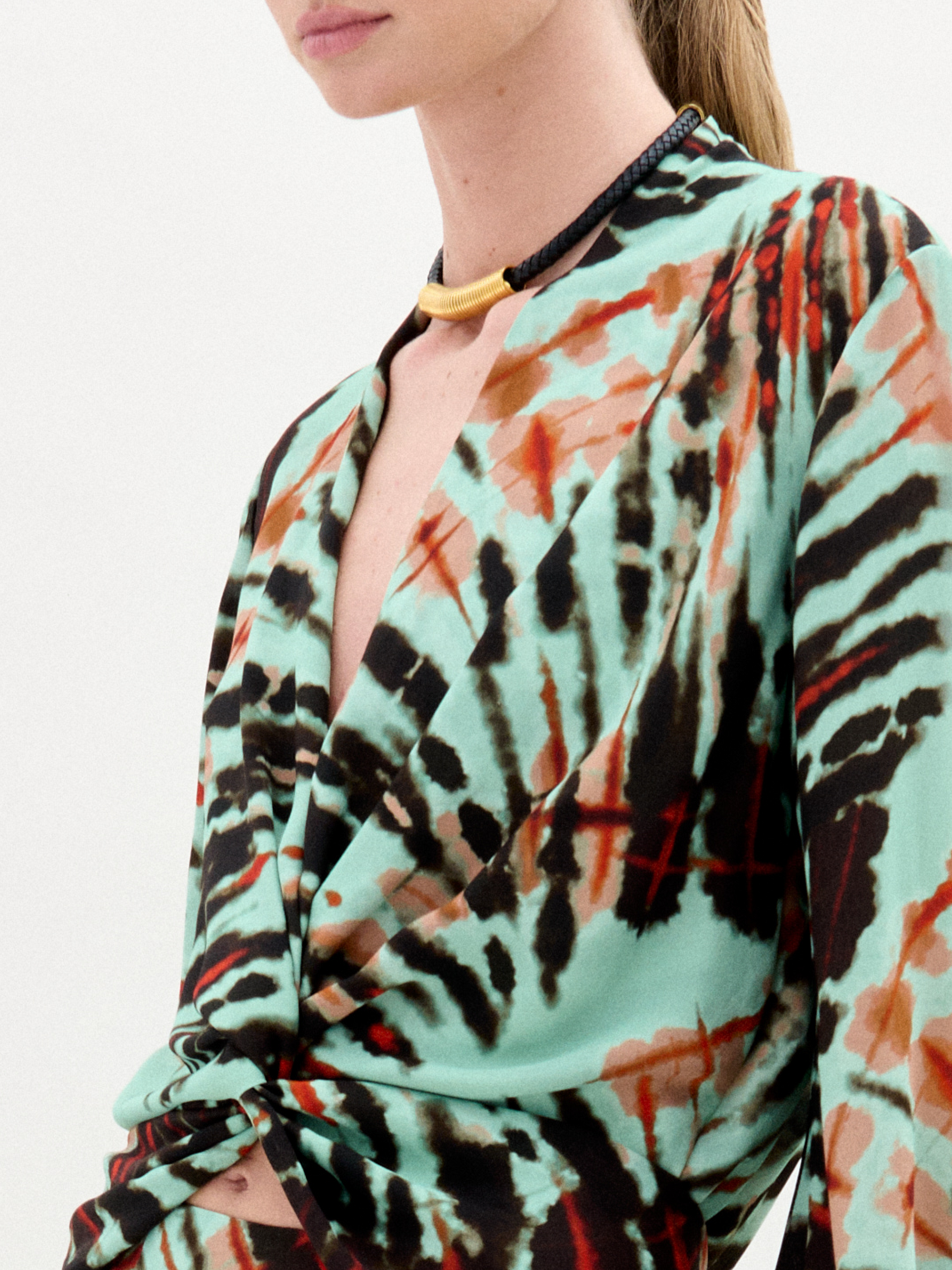 A woman stands against a plain white background wearing the Honey Blouse, a long-sleeved top featuring green, black, and orange abstract patterns with a chic front twist detail.