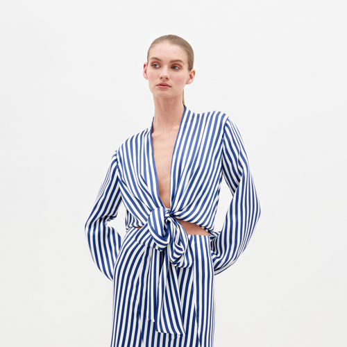 A person stands against a plain background in SVT DESIGNS' Honey Blouse, crafted from Italian textile. It features blue and white stripes, a deep V-neckline, and a waist knot tie. The individual’s hair is pulled back with hands resting on their hips.