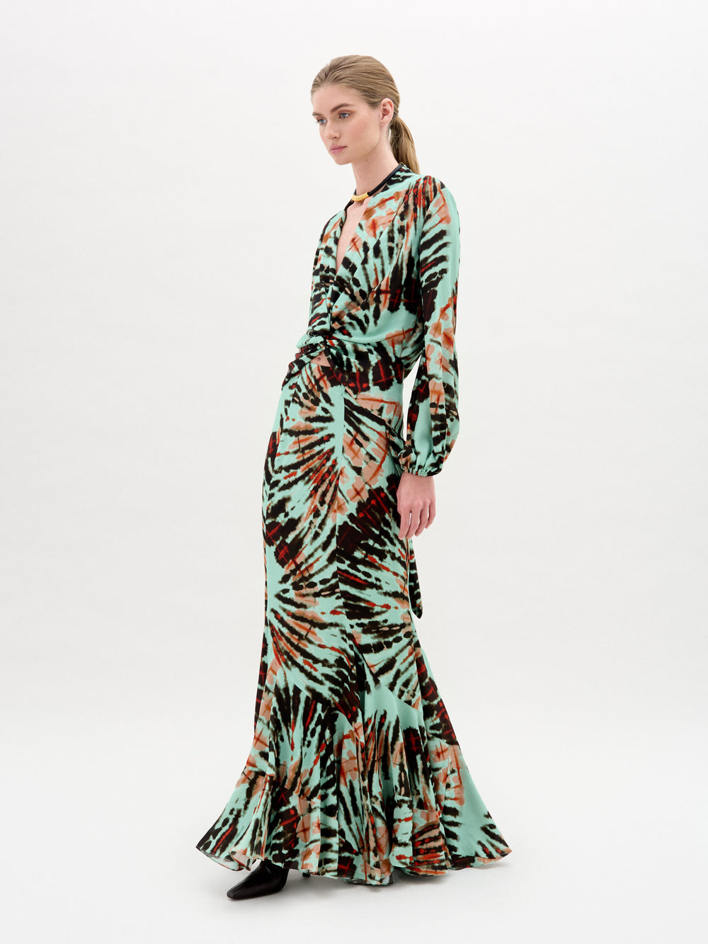 A person models the Florina Skirt by SVT DESIGNS, showcasing its long, vibrant green, black, and brown tie-dye pattern. The fitted waist accentuates a mermaid silhouette. The setting is a simple white backdrop.